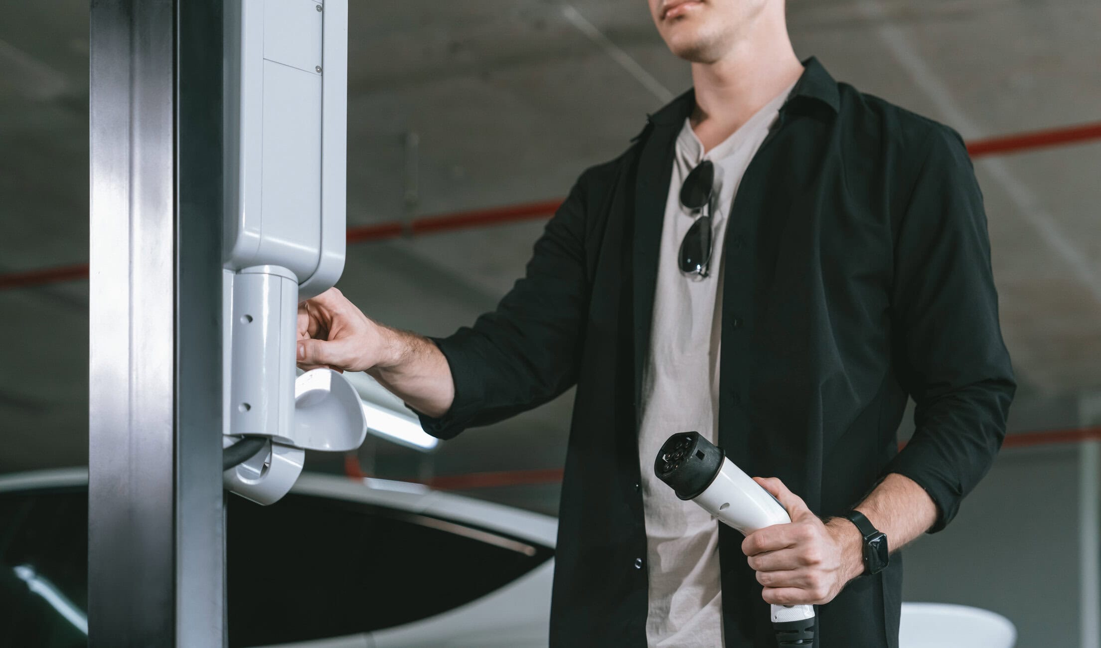 Man in a dark jacket operating a cordless drill on a white robotic arm in an industrial or parking garage setting.