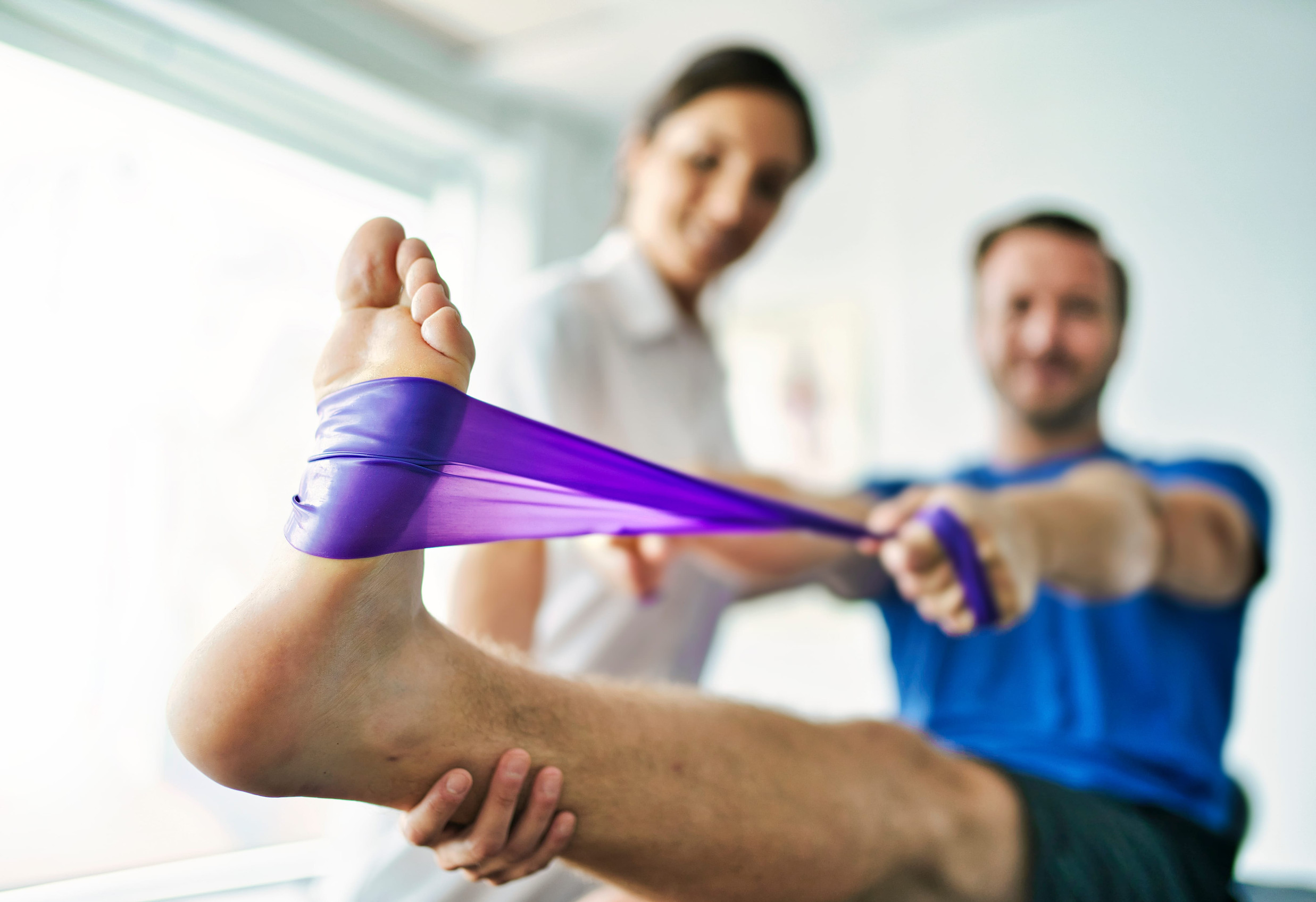 Physical therapists assist a client in a leg stretch using a purple resistance band during a physiotherapy session.
