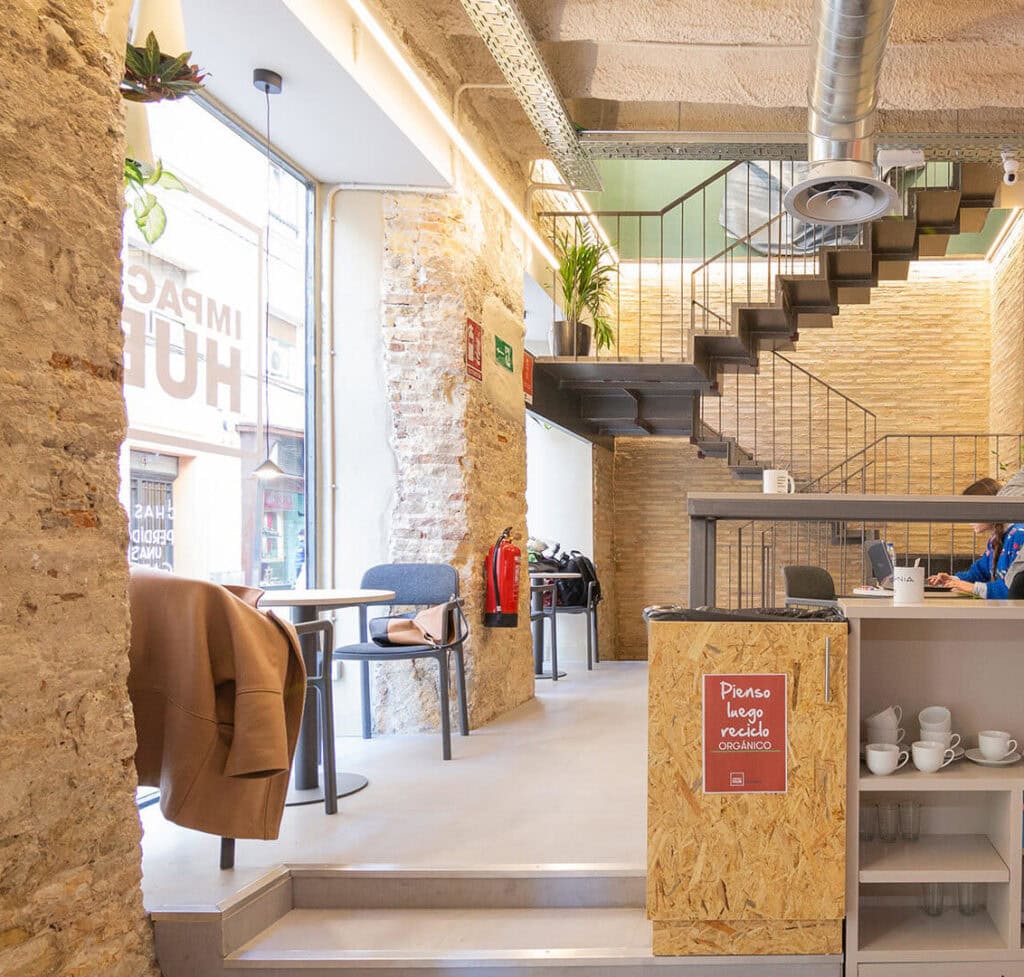 Modern cafe interior with exposed brick walls, a metal staircase, and plants near a large window.