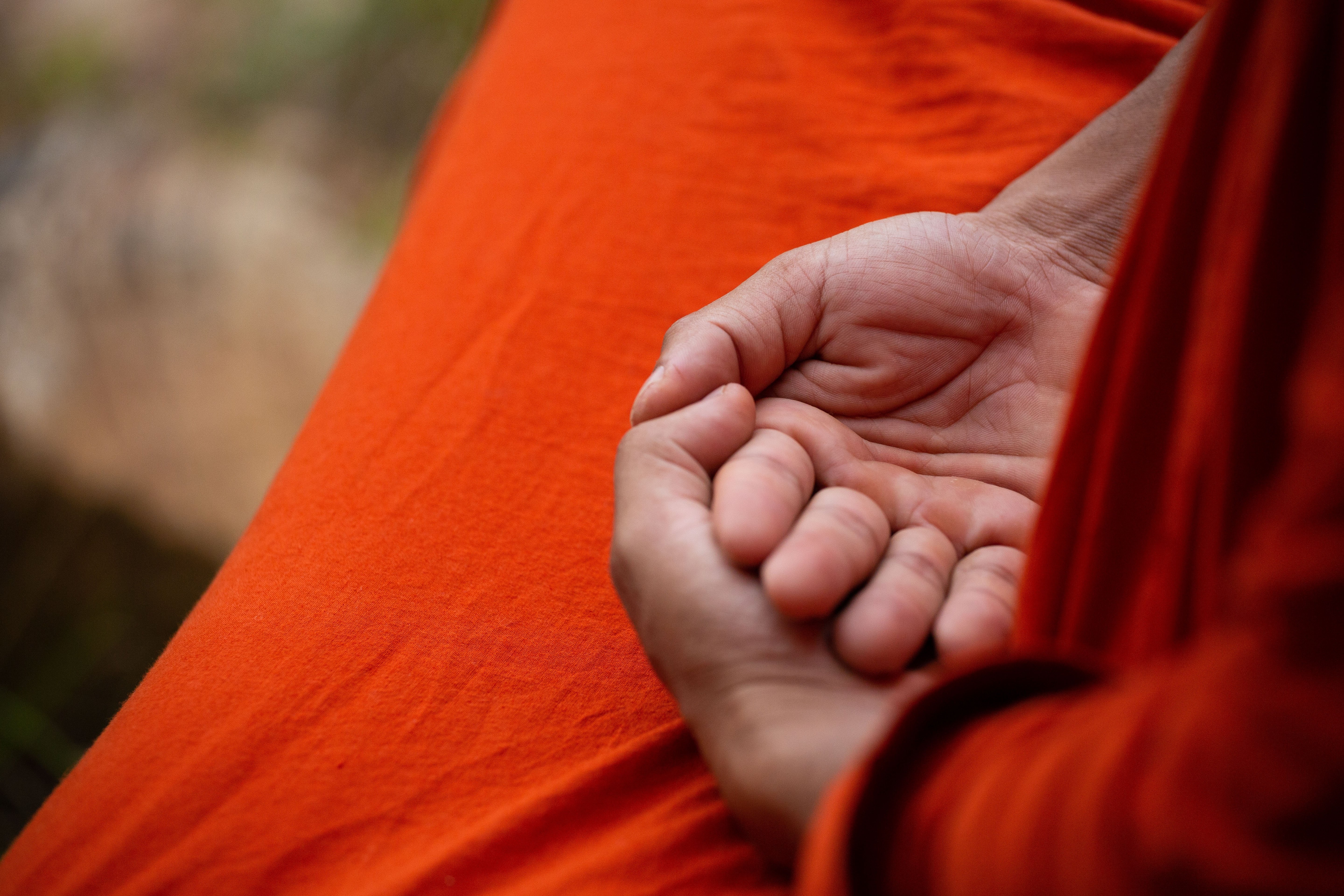 Close-up of hands folded together resting on bright orange fabric, suggesting a monk's robe maneuvered in a calm pose.
