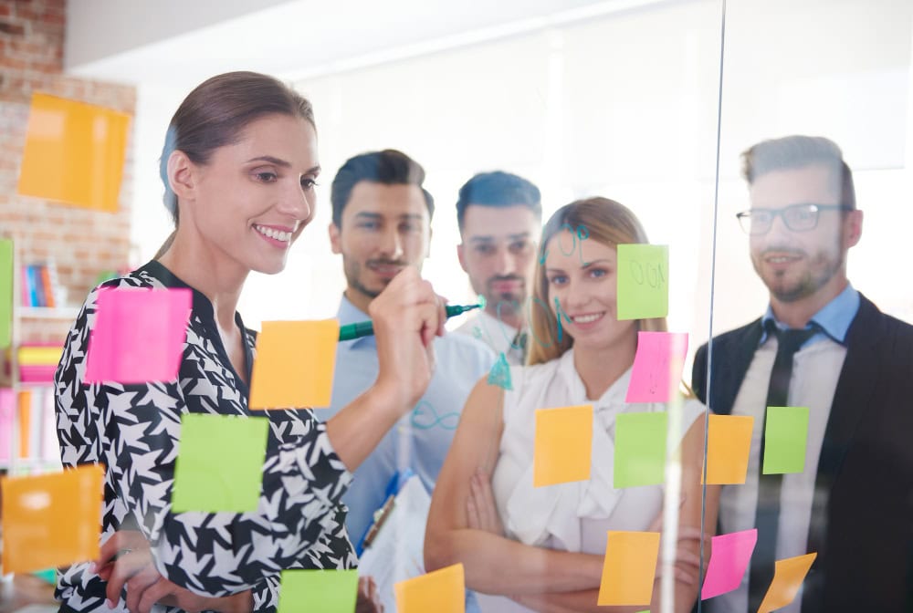 Team of five colleagues in a modern office placing colorful sticky notes on a glass wall during a brainstorming session.