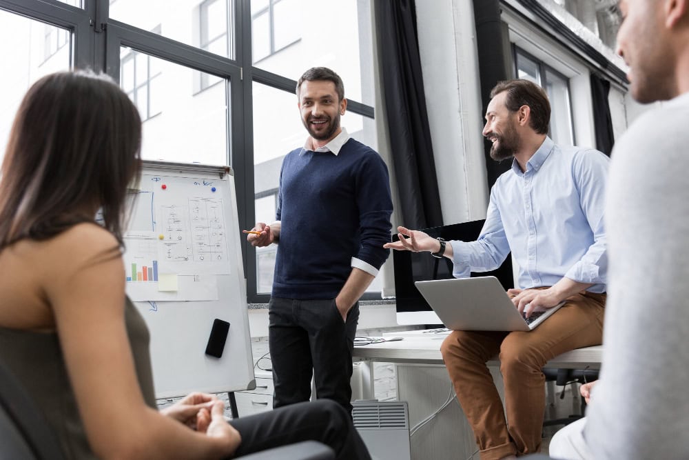 Team meeting in a bright office: man presenting with a marker beside a whiteboard as others listen attentively, laptop on the desk nearby