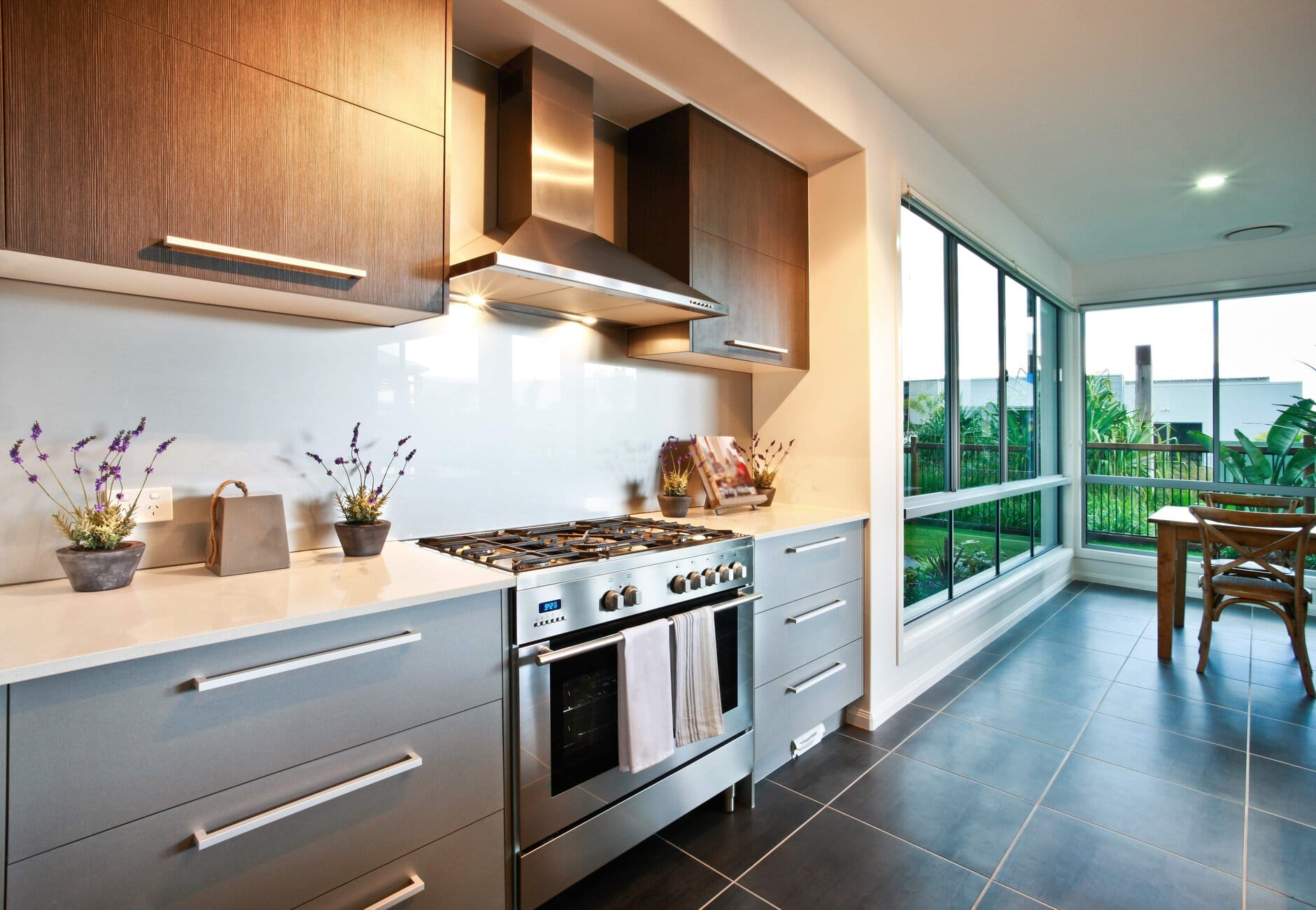 Modern kitchen with stainless steel range, hood, and dark wood cabinets along a white countertop. A bright dining nook and large windows outside.