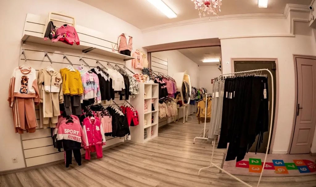 A clothing store interior with racks of kids' clothes on display and pastel walls, shelves, and a checkout area in the back.