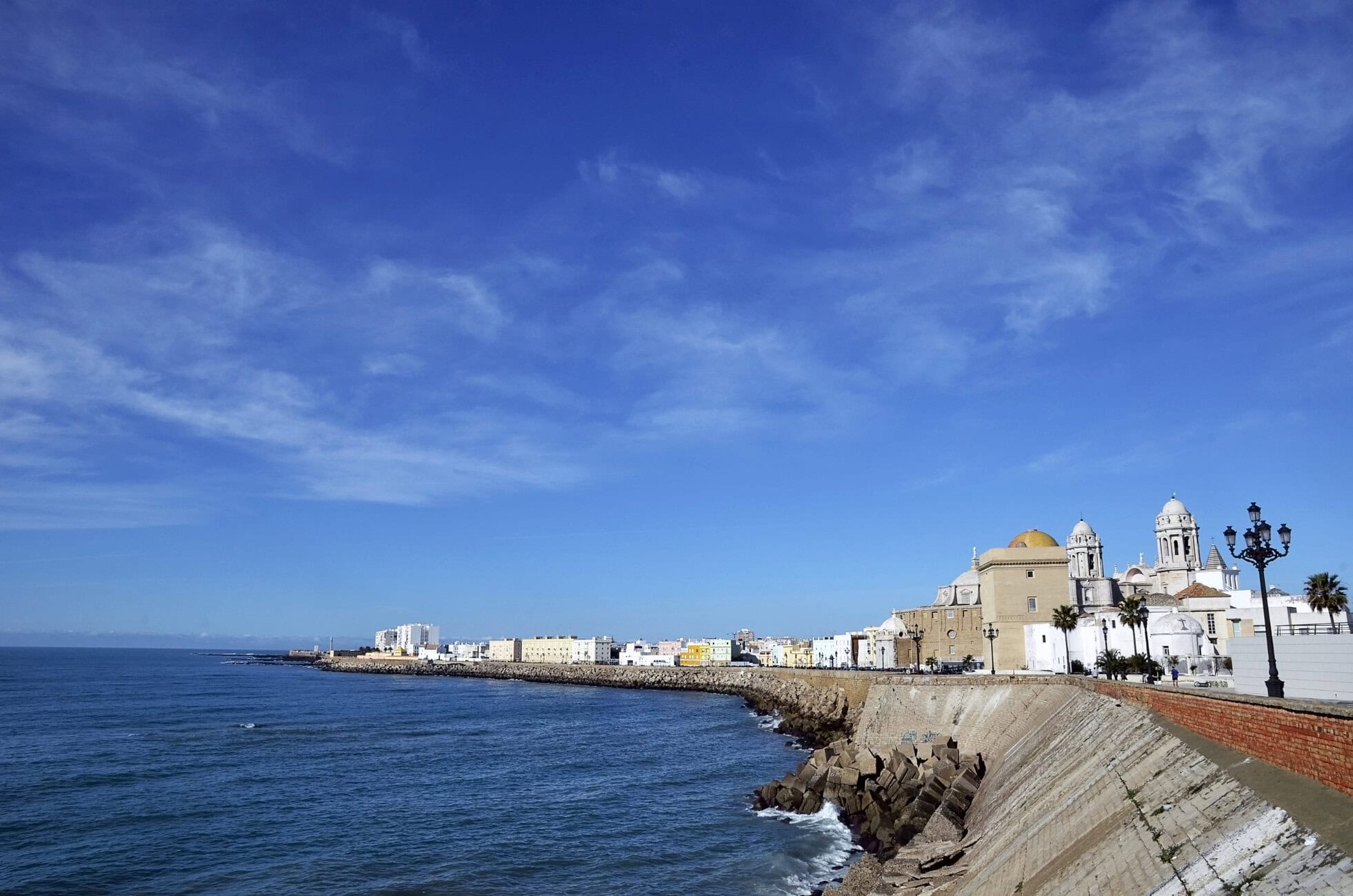 Seaside cityscape with a stone breakwater, calm blue ocean, and white domed buildings along the shore.