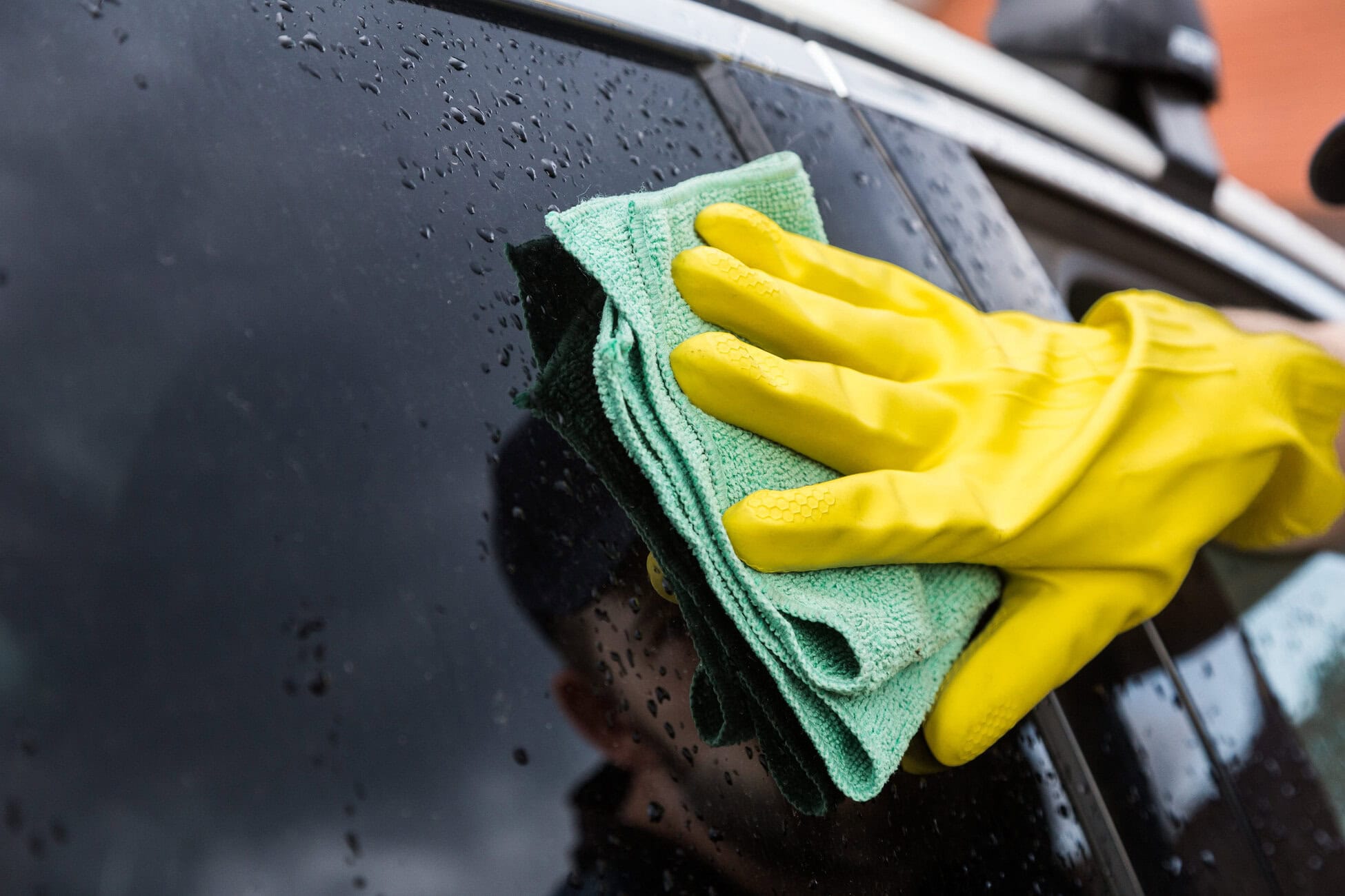 Hands in yellow gloves wiping a wet car window with a green microfiber cloth, close-up.