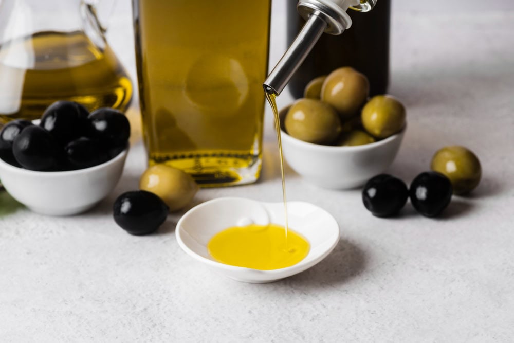 Oil is being poured from a bottle into a small white dish, with bowls of green and black olives in the background.