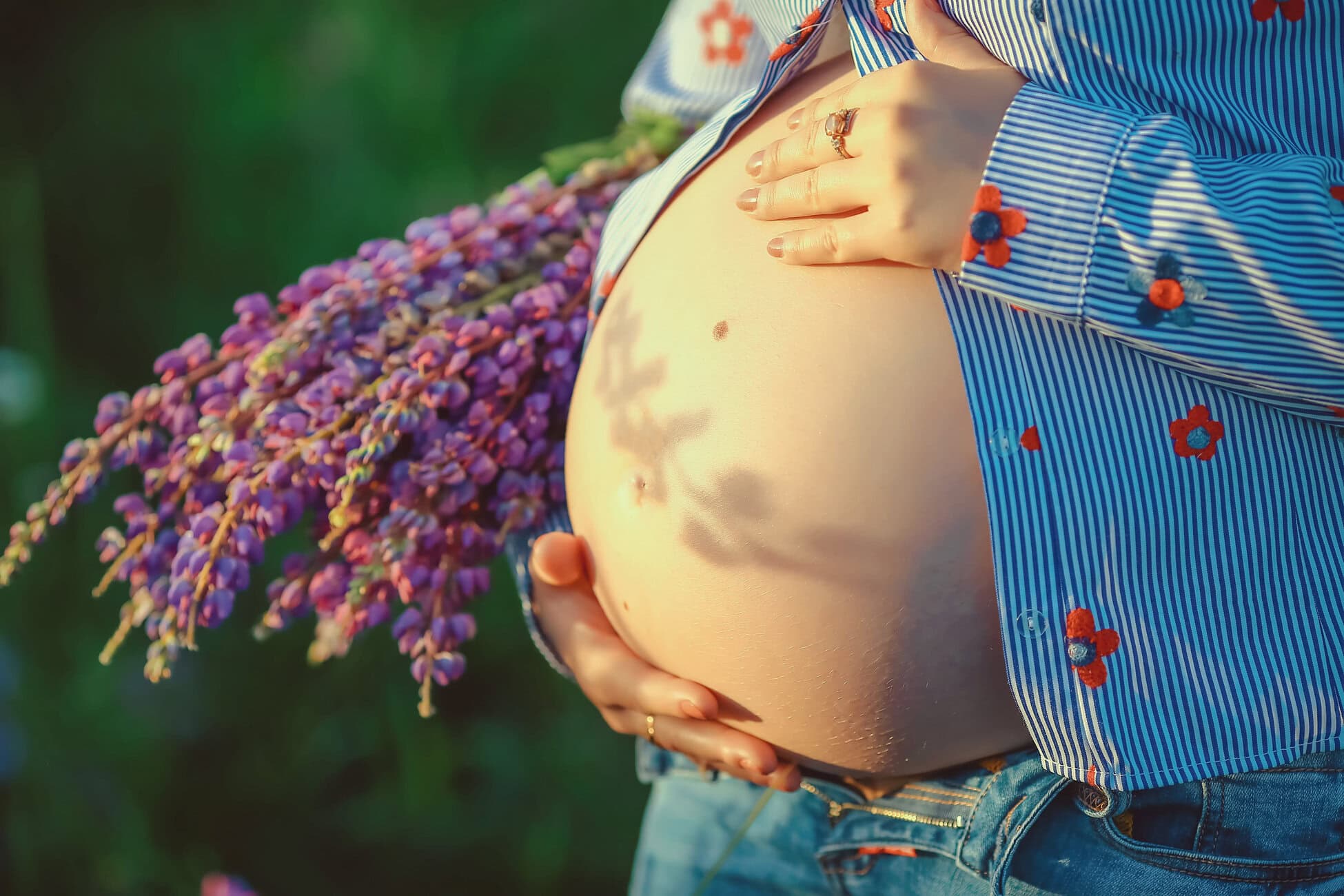 Pregnant woman gently holding her baby bump outdoors, wearing a striped shirt and jeans with purple flowers nearby.