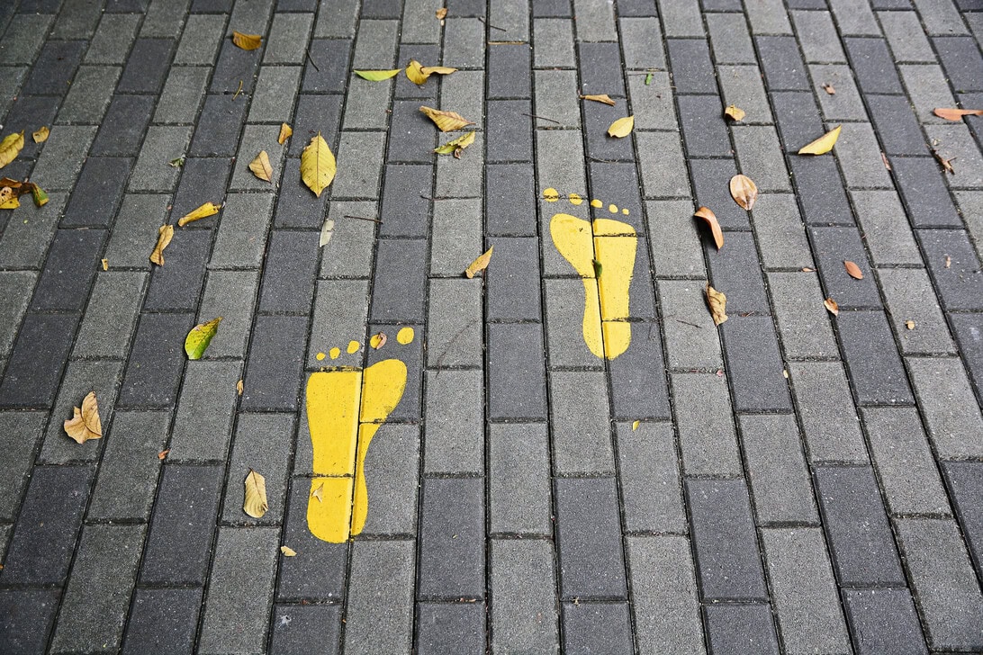 Two yellow footprint stencils on a grey brick sidewalk with scattered autumn leaves.