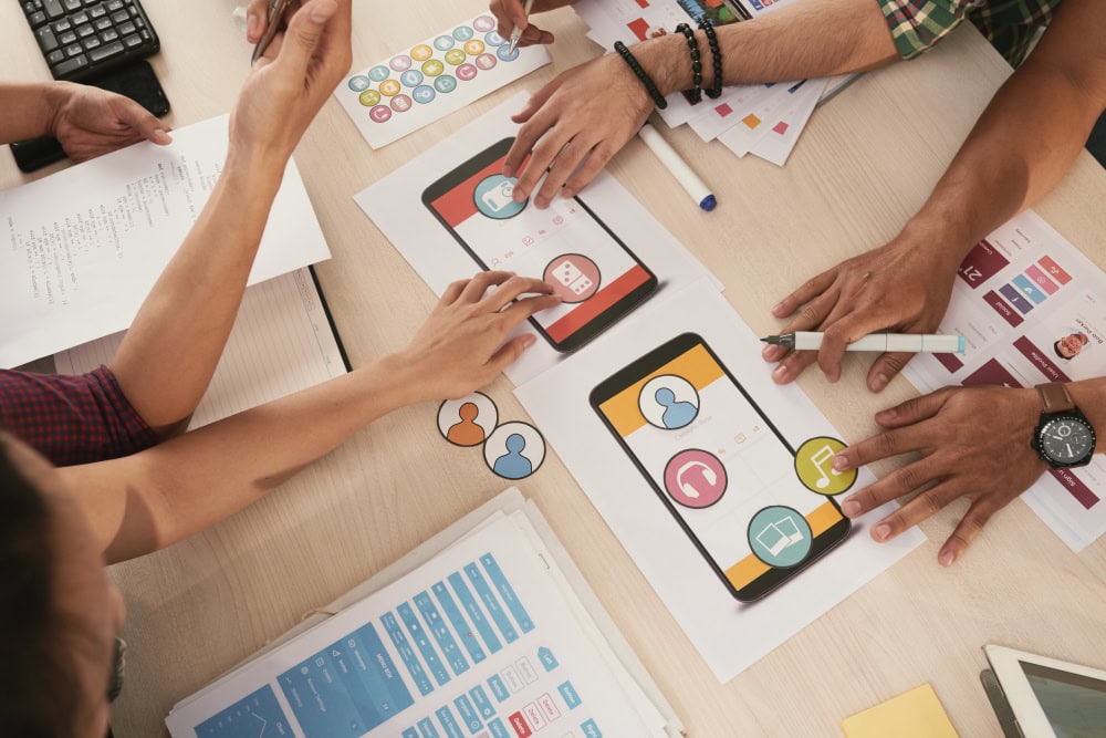 Group of people brainstorming around a table, working on mobile app mockups and charts in a design session.