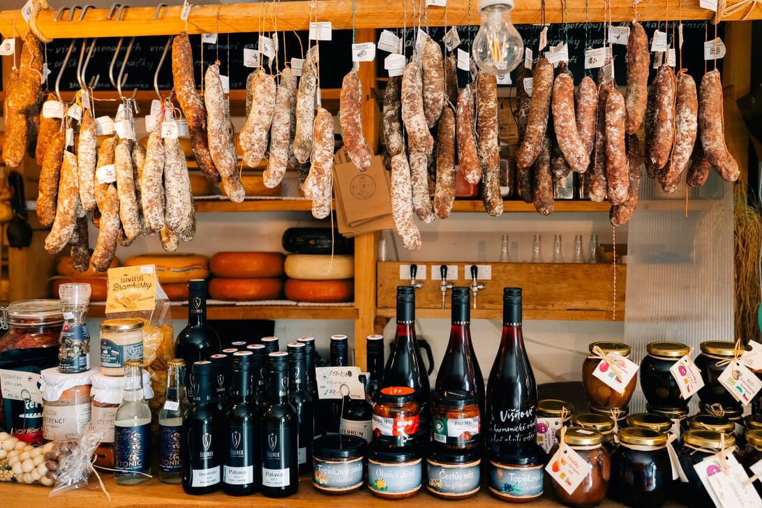 Market stall with hanging salami sausages above a display of cheeses, bottles, and jars on a wooden counter.