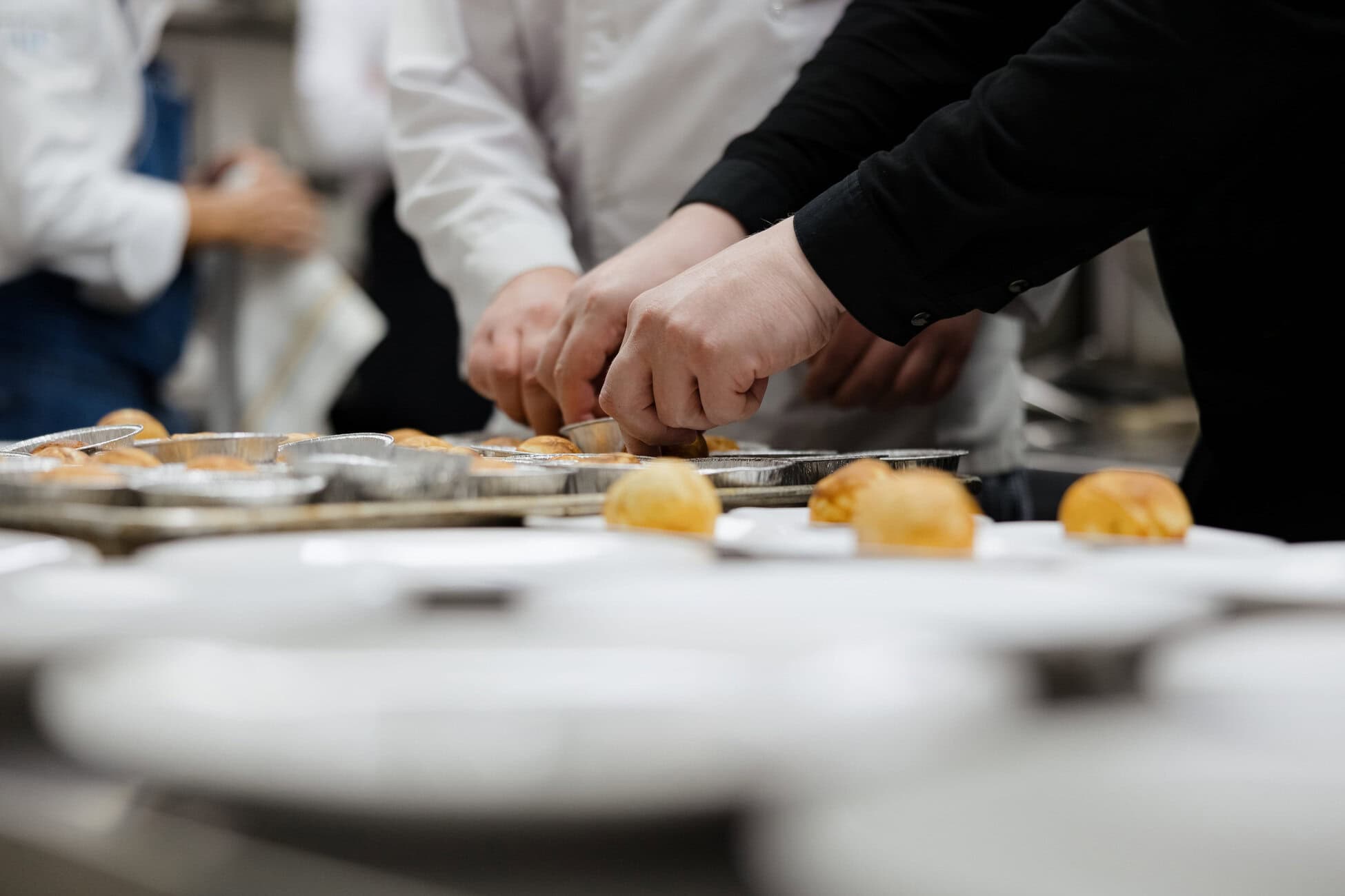 Hands of cooks shaping small pastries on metal trays in a busy kitchen.