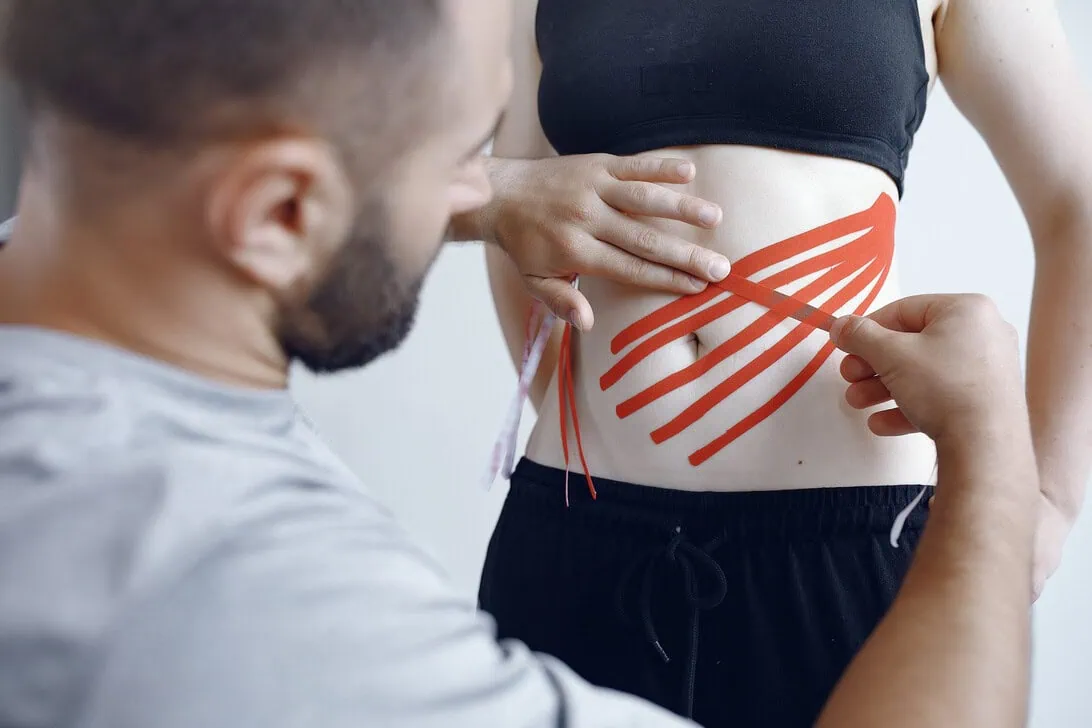 Therapist applying red kinesiology tape to a woman's abdomen in a crisscross pattern