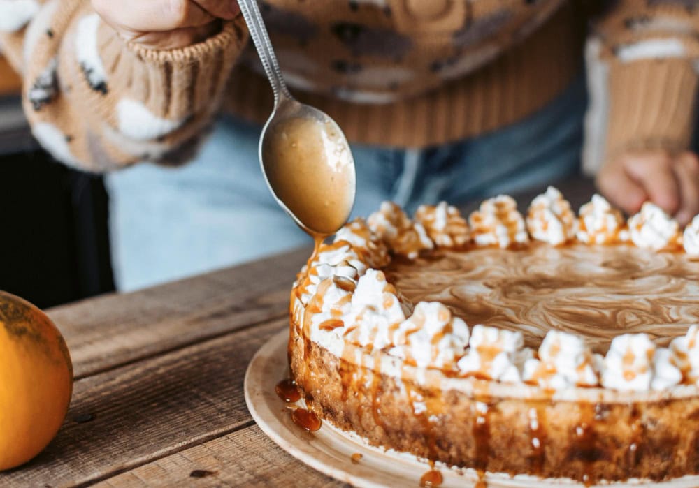 Person drizzling caramel sauce onto a caramel cheesecake topped with piped whipped cream on a wooden table, wearing a cozy sweater.