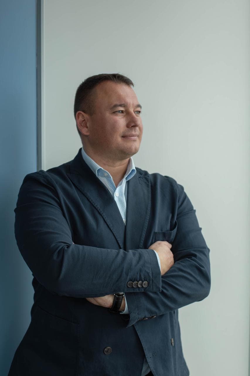 Man in a navy blazer with arms crossed, looking out a window to the right.