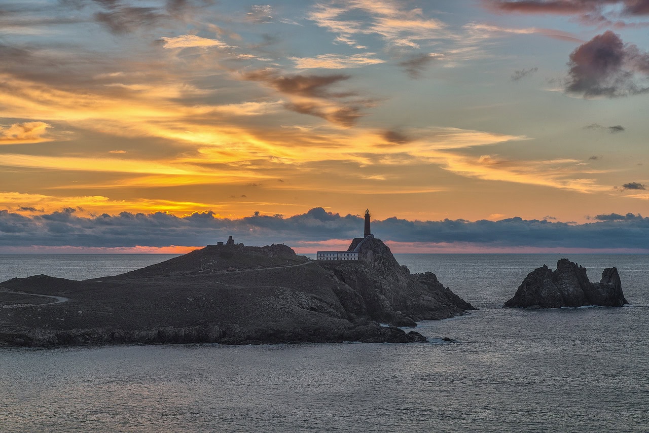 Lighthouse perched on a rocky island at sunset, with calm sea and orange-pink sky.