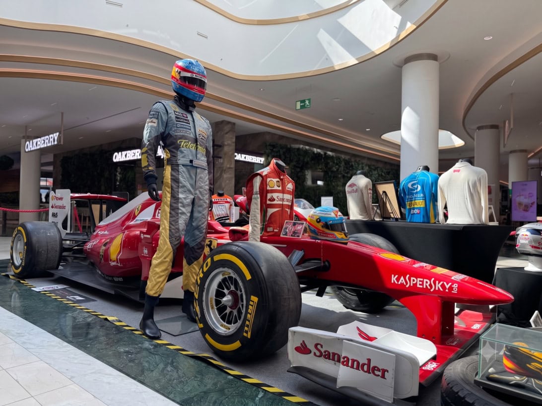 Red Formula 1 car on display in a mall, with a racer mannequin in a gray and yellow suit standing beside it and a racing helmet on the hood.