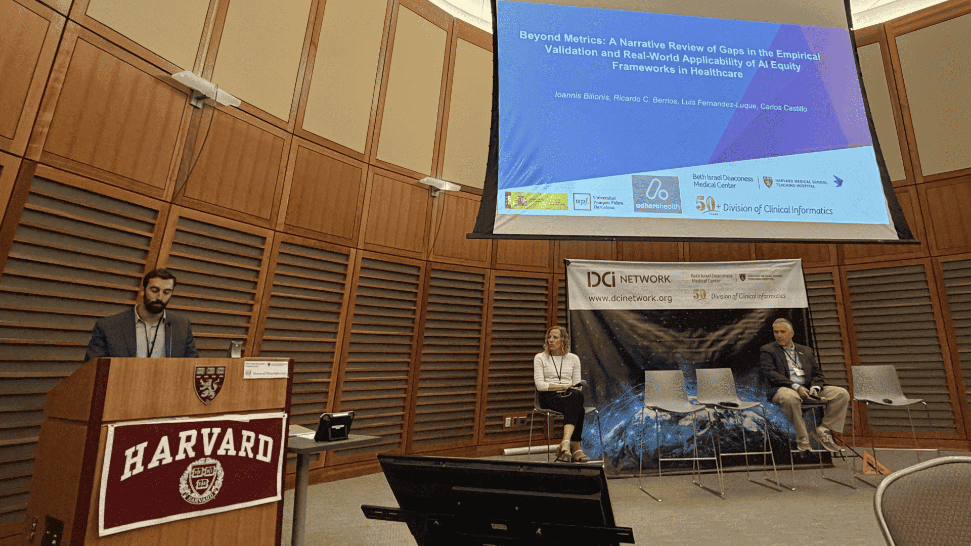 Speaker at a Harvard podium delivering a talk, with a large projection screen overhead and two panelists seated on stage to the right.