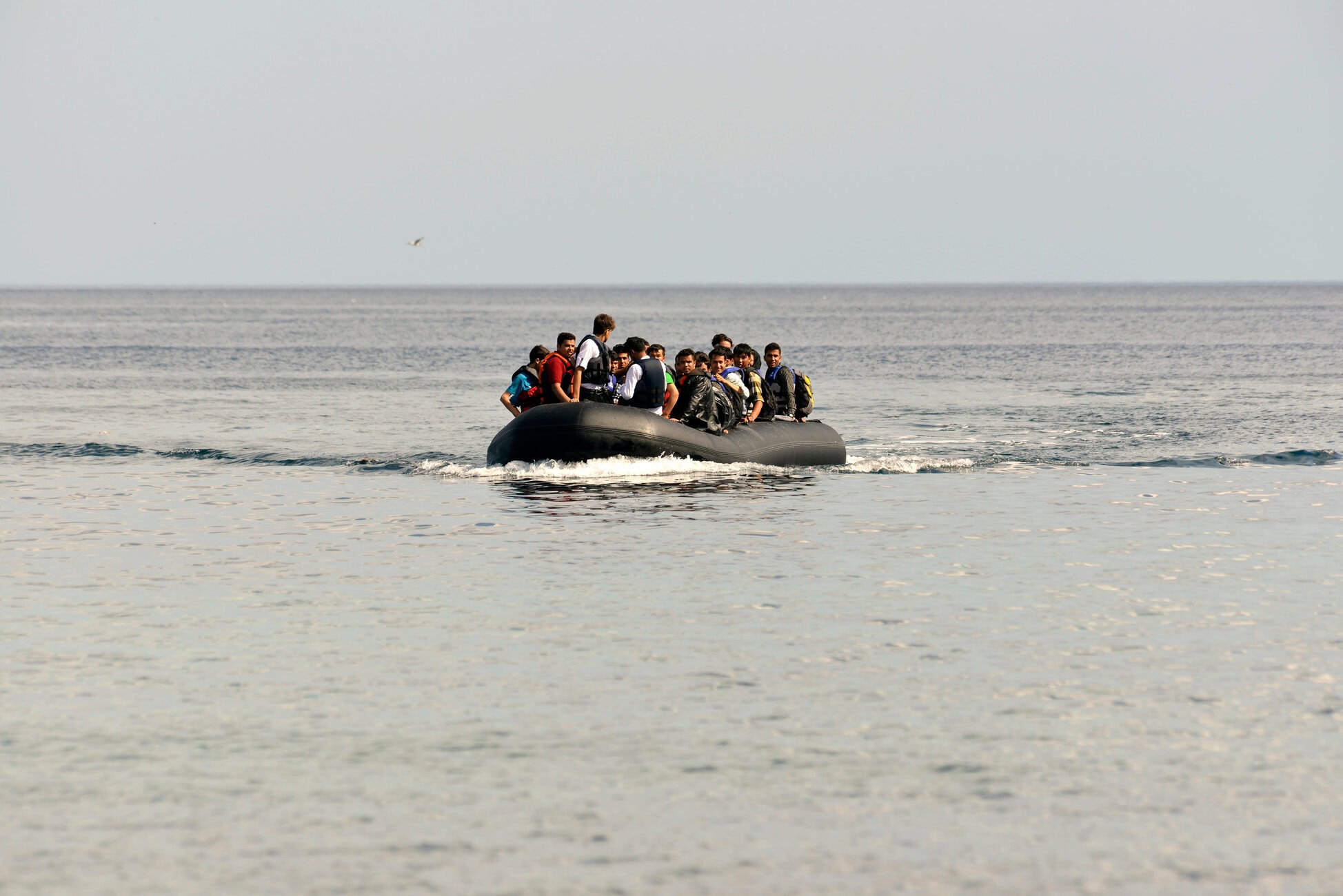 A group of people wearing life jackets crowded onto a black inflatable boat in the open sea, waves brushing the hull.