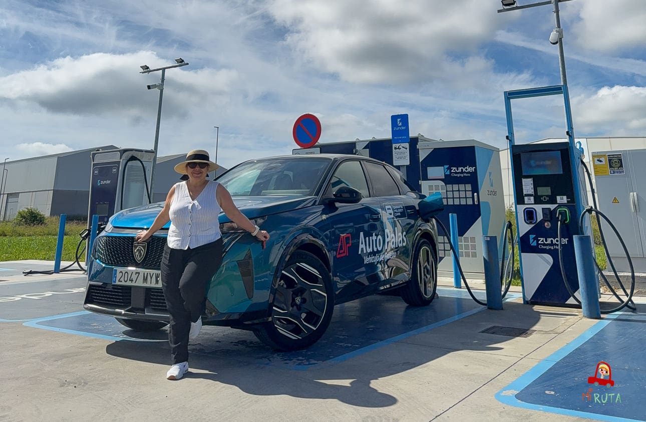 Woman wearing a straw hat and white sleeveless top leaning on a blue AutoPalas electric SUV at a public charging station.