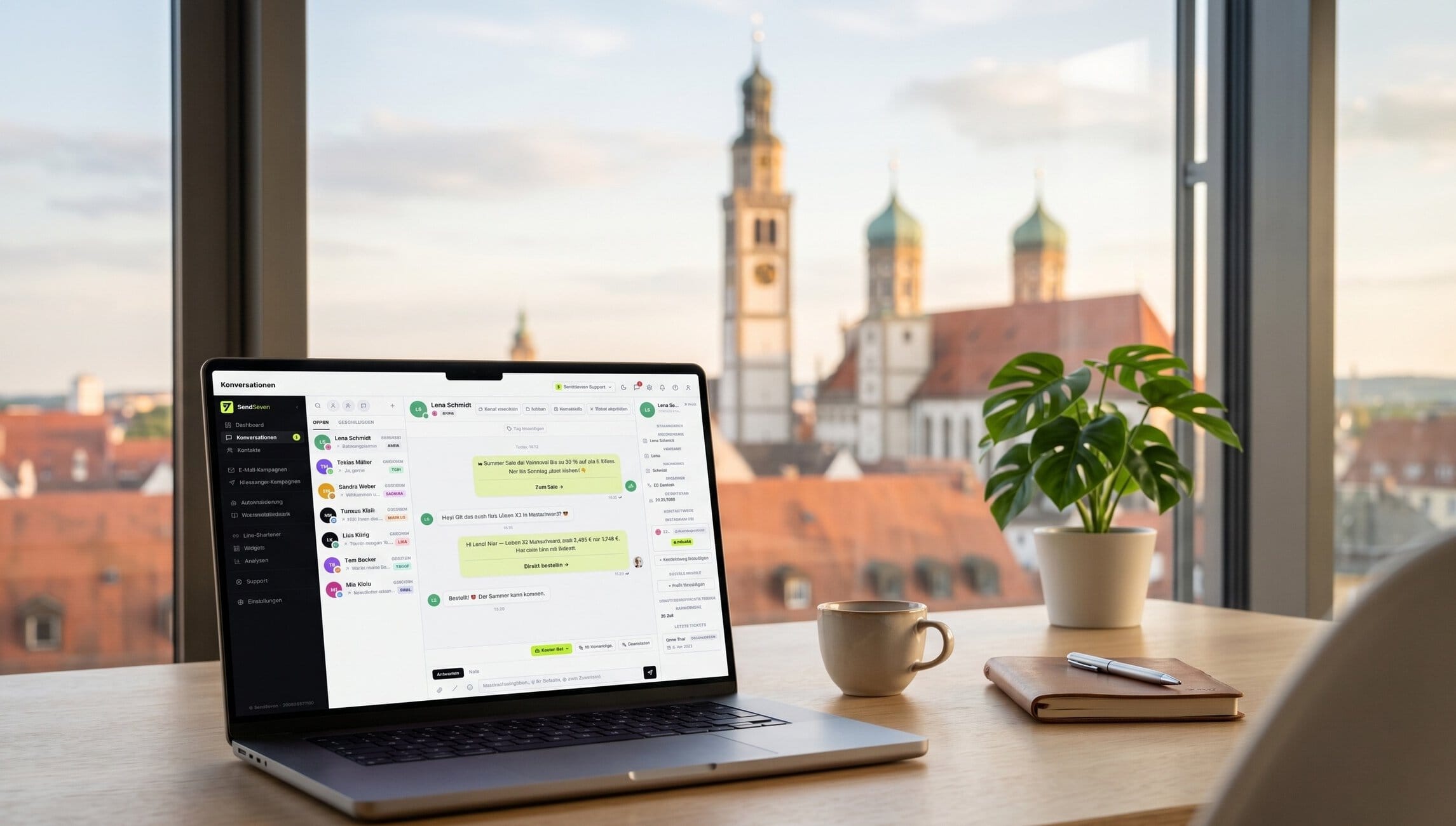 Laptop on a wooden desk shows a chat interface, with a potted plant, a coffee mug, and a notebook nearby, city buildings visible through the window.