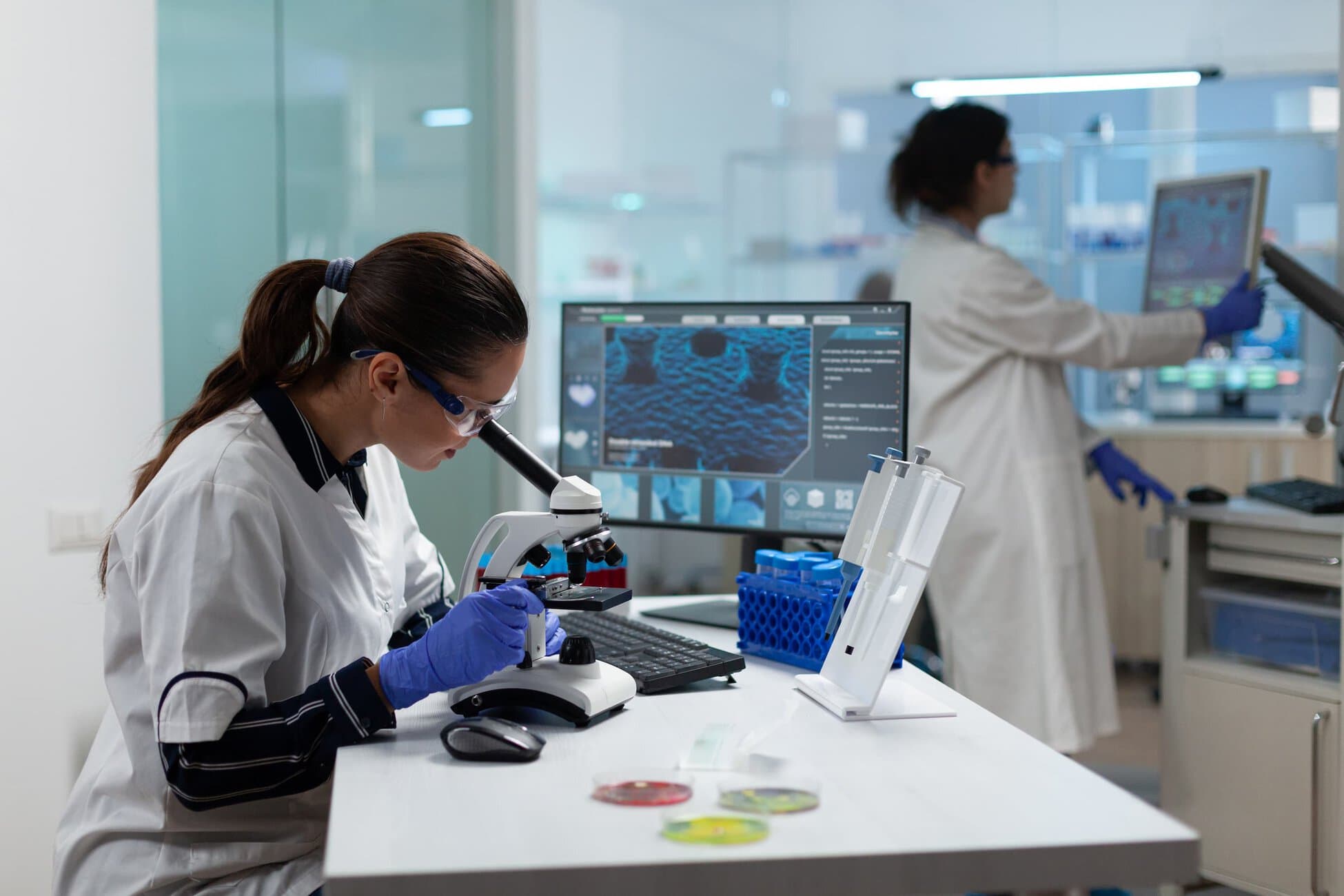 Scientist in lab coat and safety goggles examines a microscope at a bench with a computer monitor nearby in a modern lab.