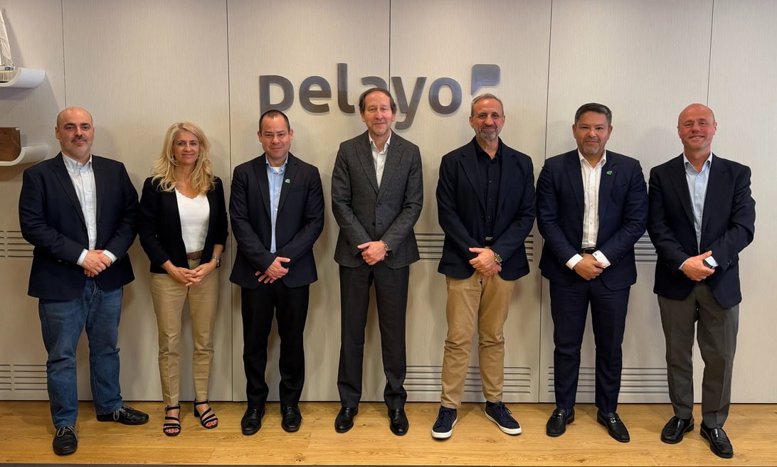 Seven professionals (six men and one woman) stand in a line in a modern office, posing in front of a wall with a company logo behind them.