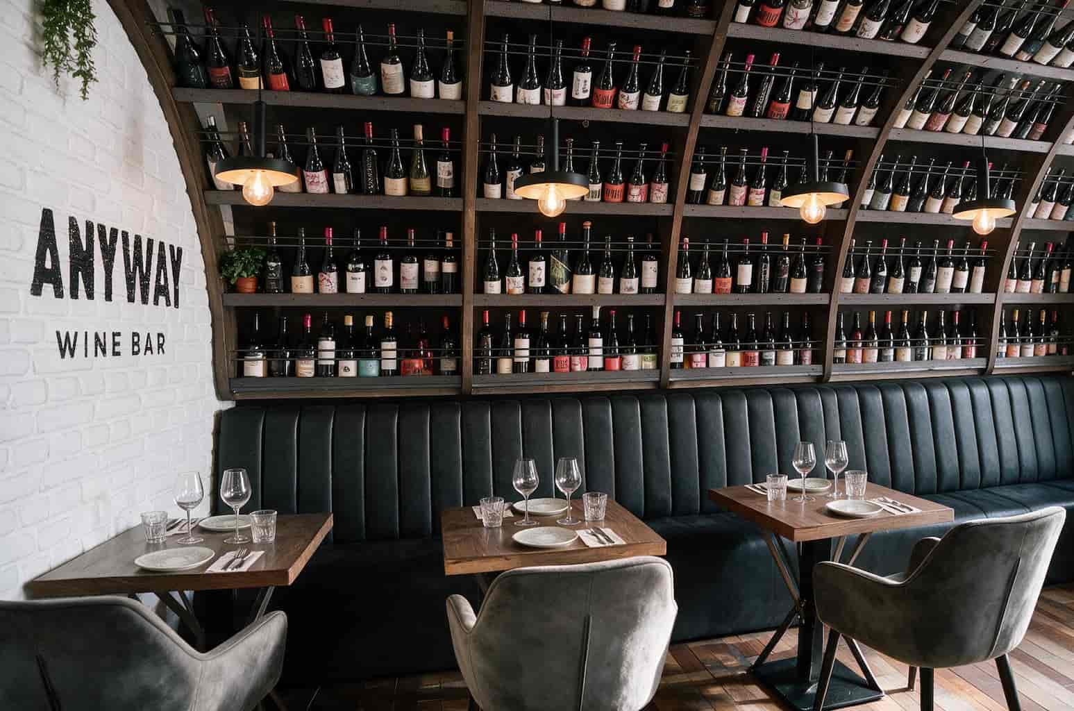 Wine bar interior with white brick wall, wall shelves of wine bottles, and a dark green upholstered banquette; tables set for guests.