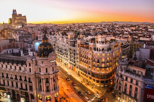 Sunset over a European city with ornate buildings and a prominent domed tower at a busy street corner, likely the Gran Via in Madrid, bathed in warm orange and pink hues.
