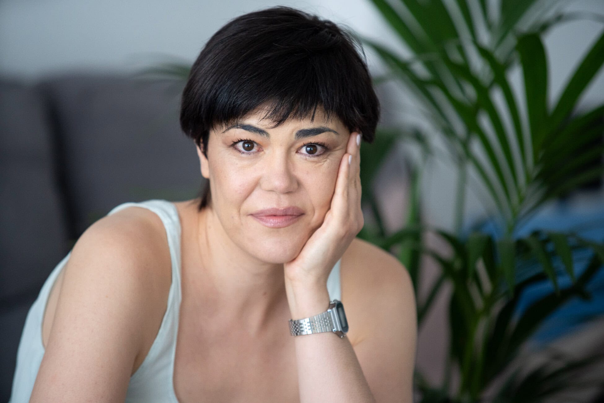 Woman with short dark hair resting her face on her hand, looking at the camera indoors with green plants in the background