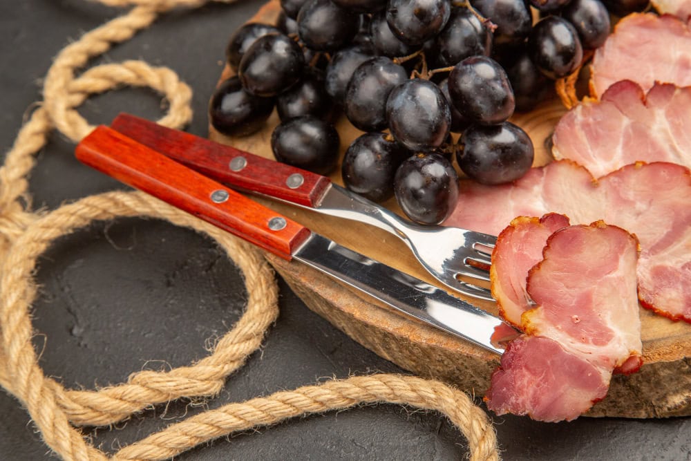 Charcuterie board with sliced bacon, dark grapes, and forks on a wooden slab; rope decor visible nearby.
