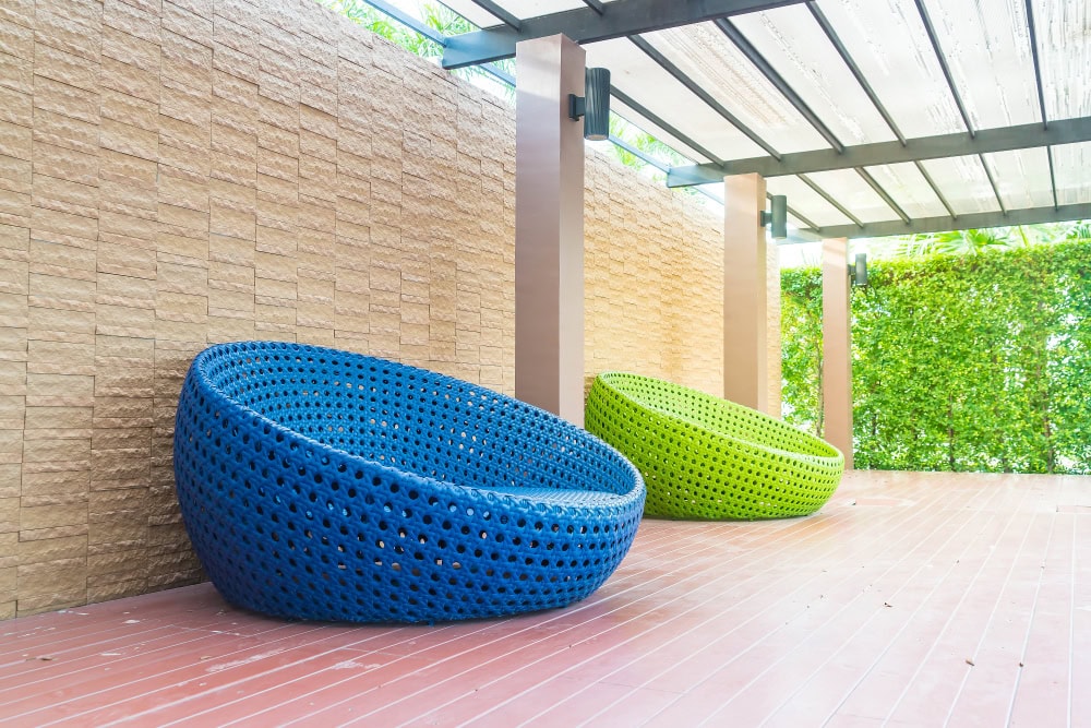 Blue and green round wicker lounge chairs sit on a wooden deck under a glass-roofed patio by a beige brick wall. (Informative, concise)