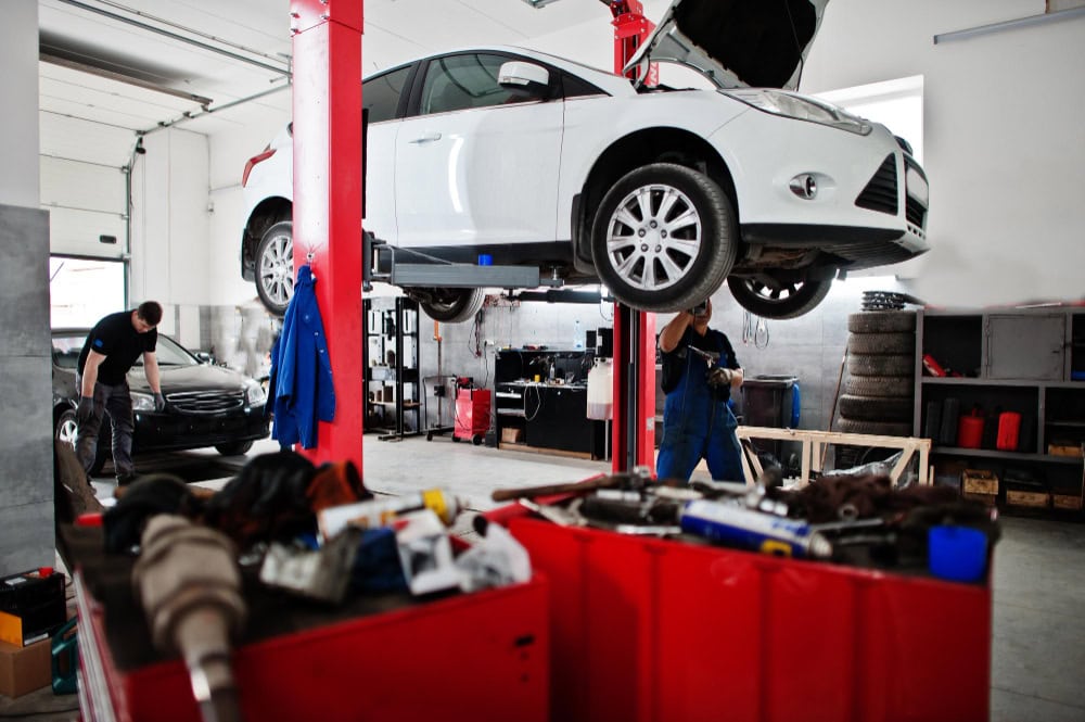 White car lifted on a hydraulic lift in an auto repair shop, two technicians inspecting the undercarriage, red tool chest in the foreground.