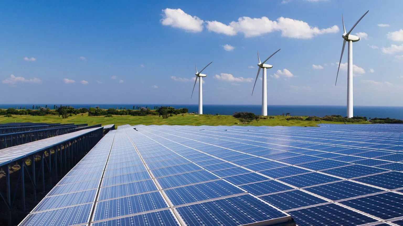 Rows of solar panels stretch toward the horizon with three wind turbines near the coast under a clear blue sky, sea in the distance.