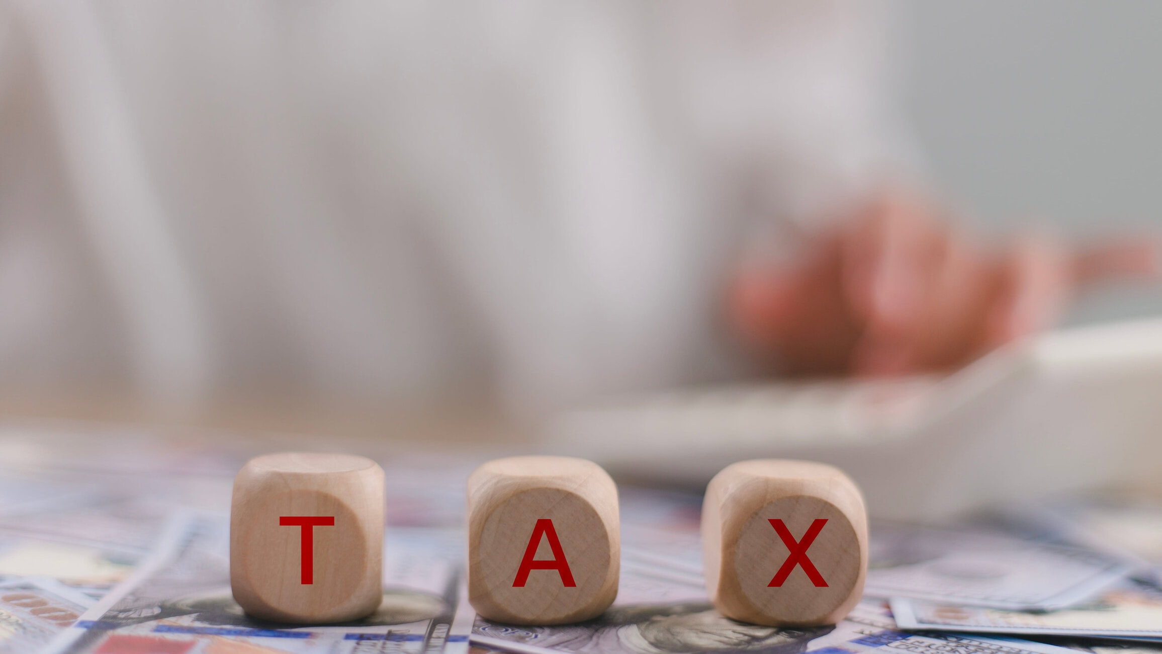 Close-up of wooden blocks spelling TAX on top of scattered banknotes with a blurred background, suggesting tax-themed imagery.