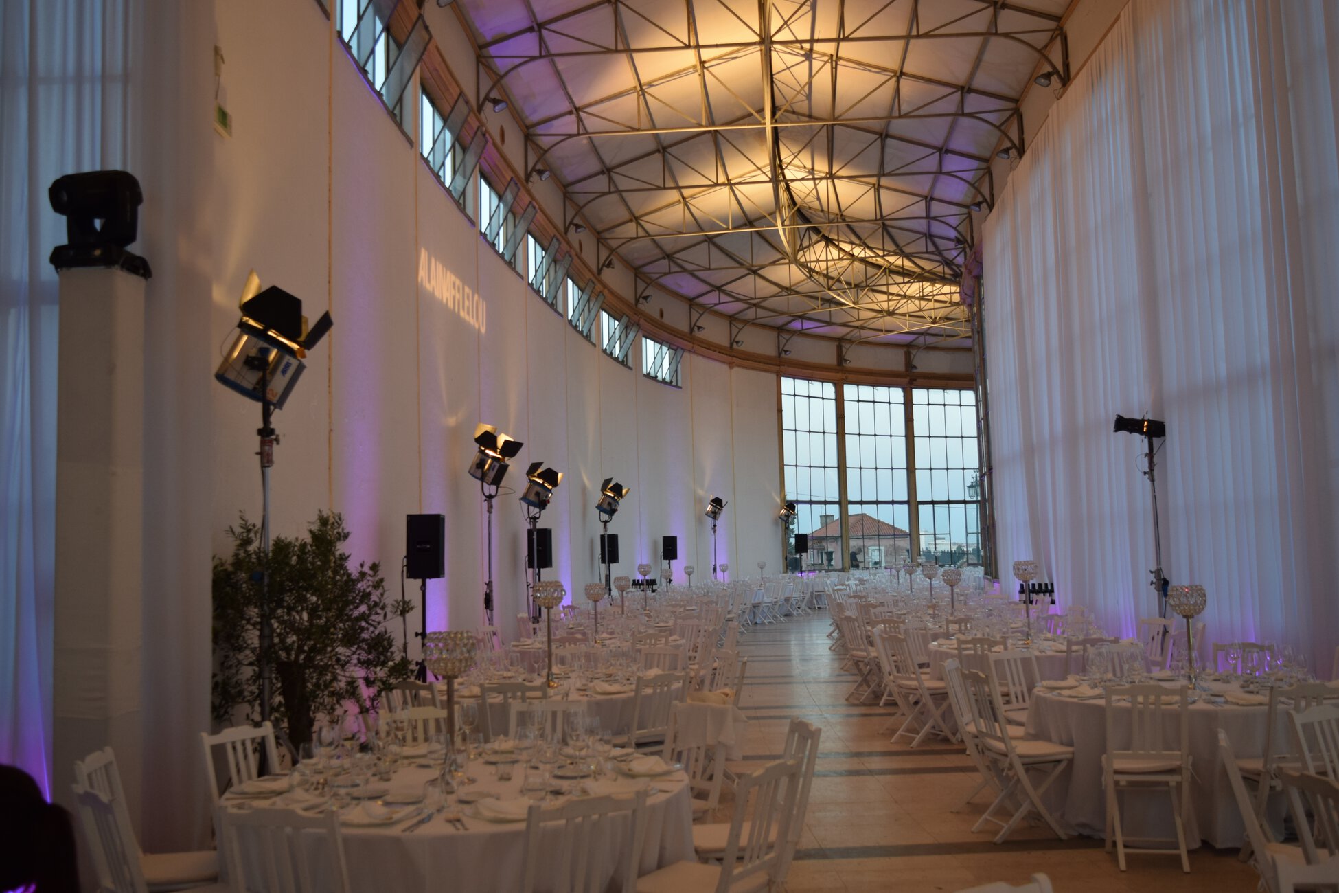 Indoor banquet hall set for a formal event with round tables, white chairs, glassware, and purple uplighting.