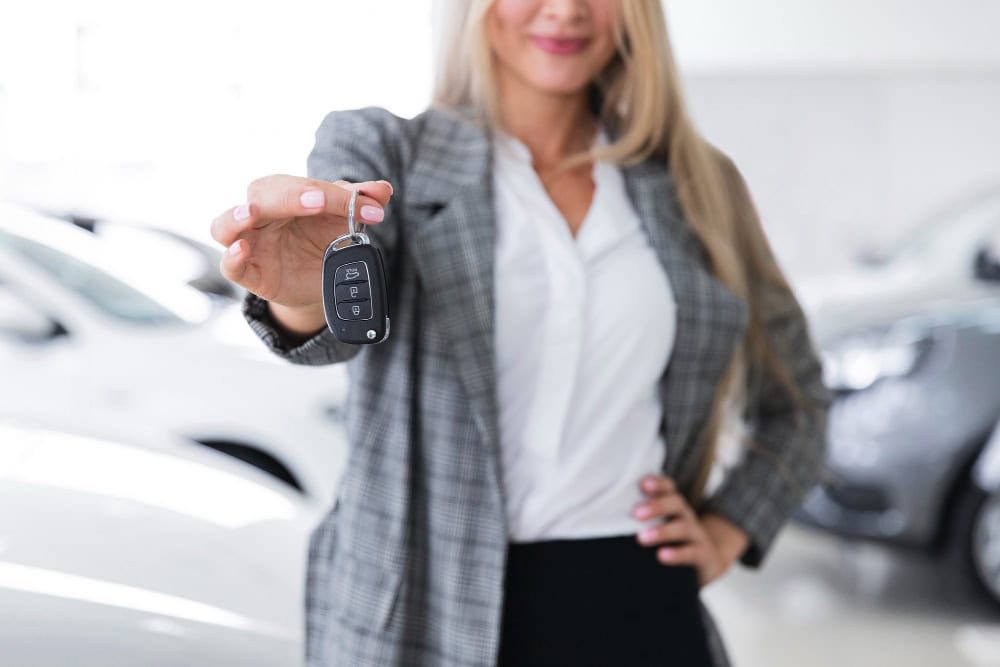 Woman in a blazer extends a car key fob toward the camera in a showroom setting, with cars blurred in the background