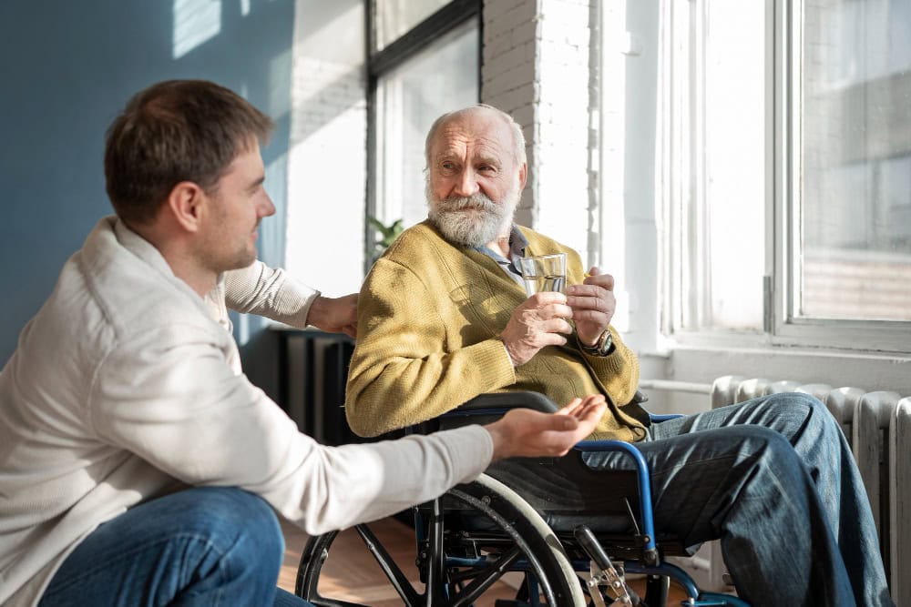 Younger man in a white sweater talks with an elderly man in a wheelchair, sharing a moment with a glass in hand, by a sunlit window.