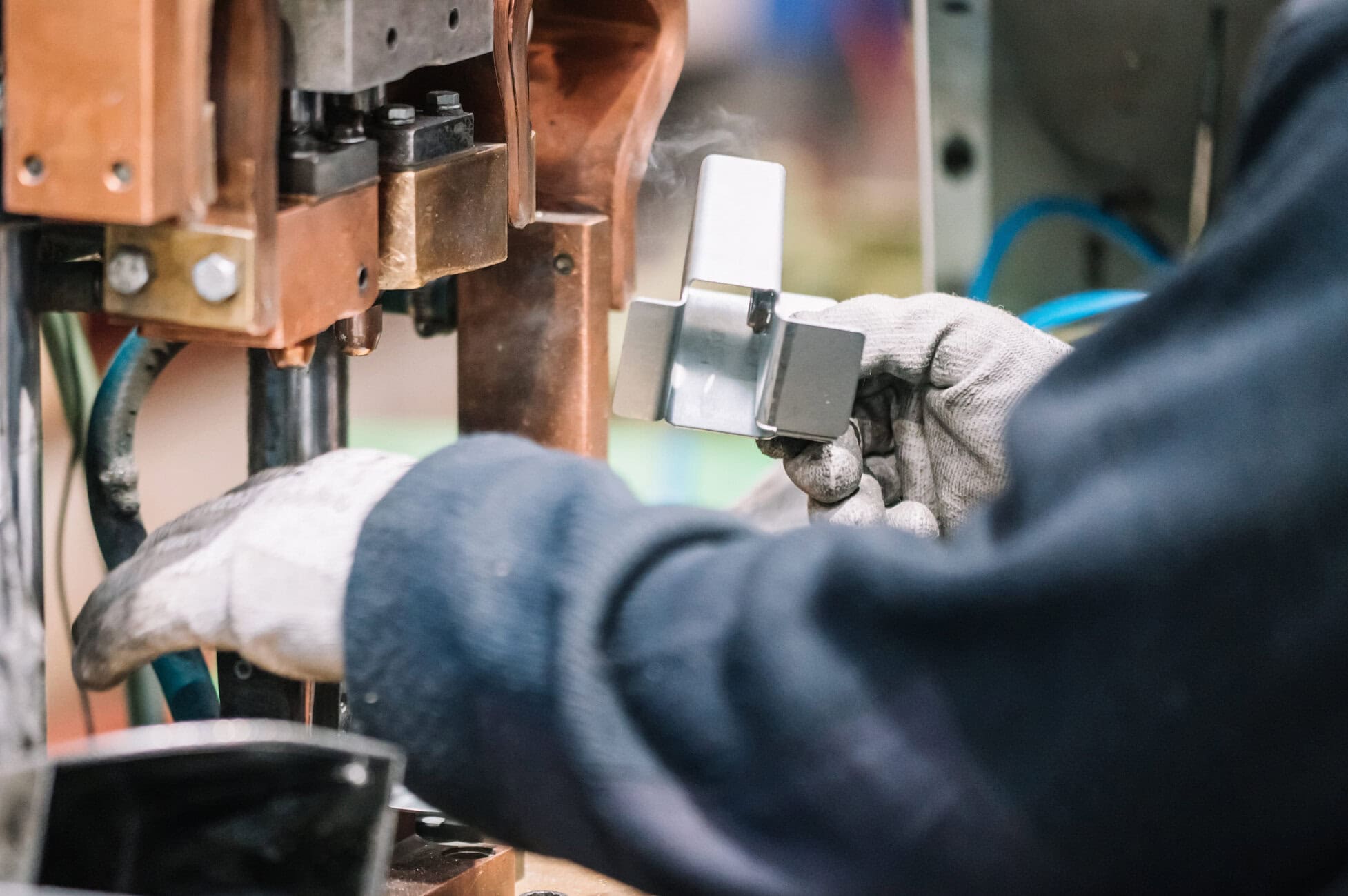 Gloved hands hold a metal piece near a copper industrial press or clamp in a workshop setting.