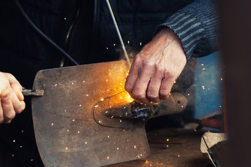 Hands of a worker shaping hot metal on a flat plate, sparks flying from the contact point on a workshop bench.