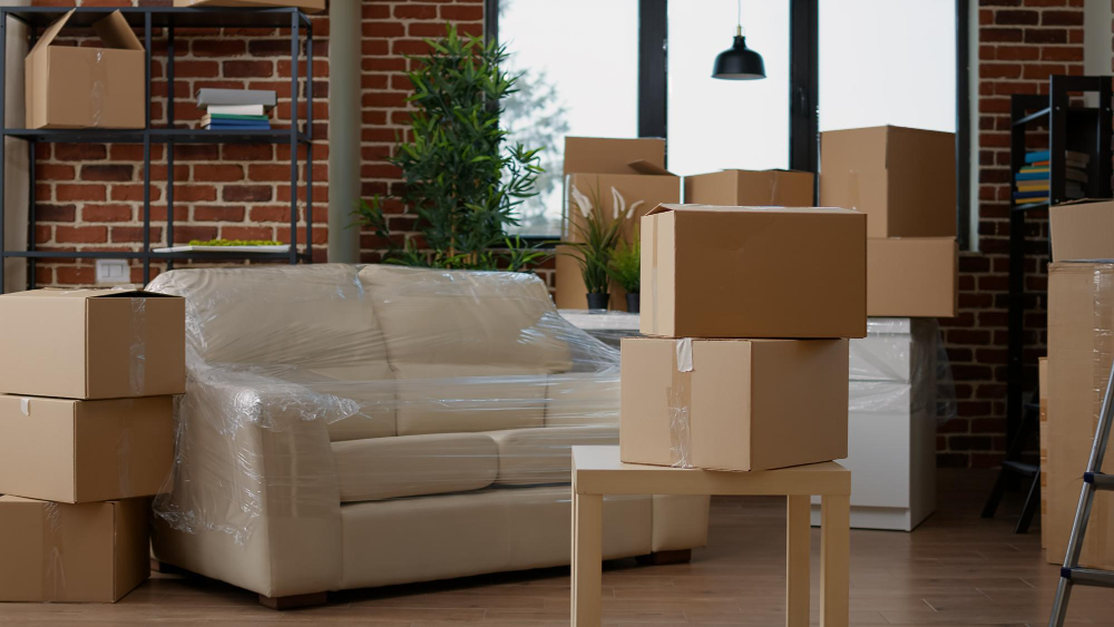 Cardboard boxes stacked on a small table and around a wrapped sofa in a brick-walled living room, suggesting packing or moving to another space.