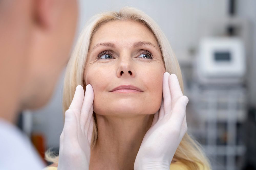 Woman undergoing a facial examination, with gloved hands supporting her jaw in a clinical setting.