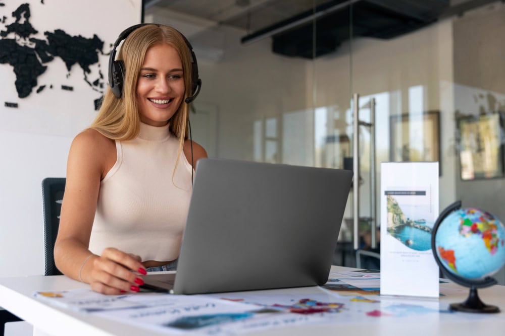 Woman with a headset smiling at a laptop on a bright desk with a globe and travel brochures nearby.