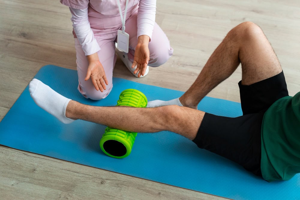 Physical therapist kneels beside a blue mat, guiding a leg rehabilitation exercise with a green foam roller under the shin.
