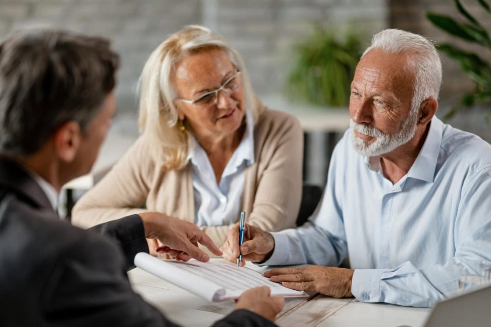 Older man with white hair signs a document at a table as a colleague points to the pages and a woman watches nearby.