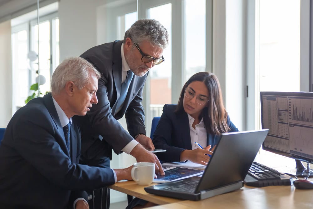 Three business professionals collaborate at a desk, examining papers and a laptop in a bright office.