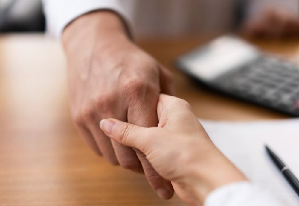 Close-up of a handshake between two professionals over a wooden desk, with a pen nearby on the desk.