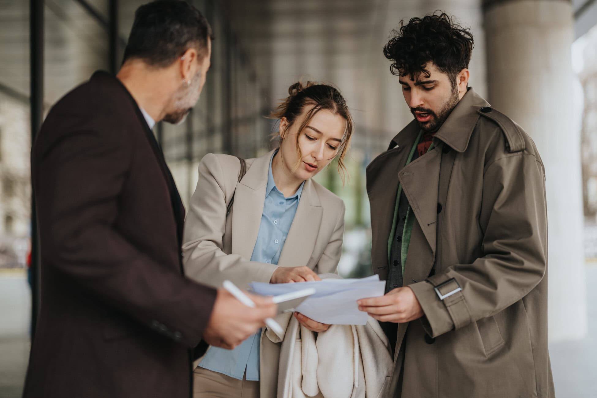 Three adults in business attire review papers outdoors beside a building. (informative)