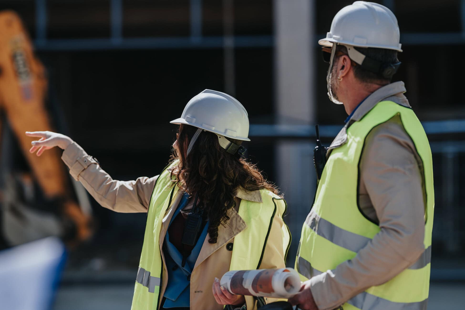 Two construction workers in hard hats and high-visibility vests discuss plans at a building site, one gesturing toward something off-camera, the other holding blueprints.