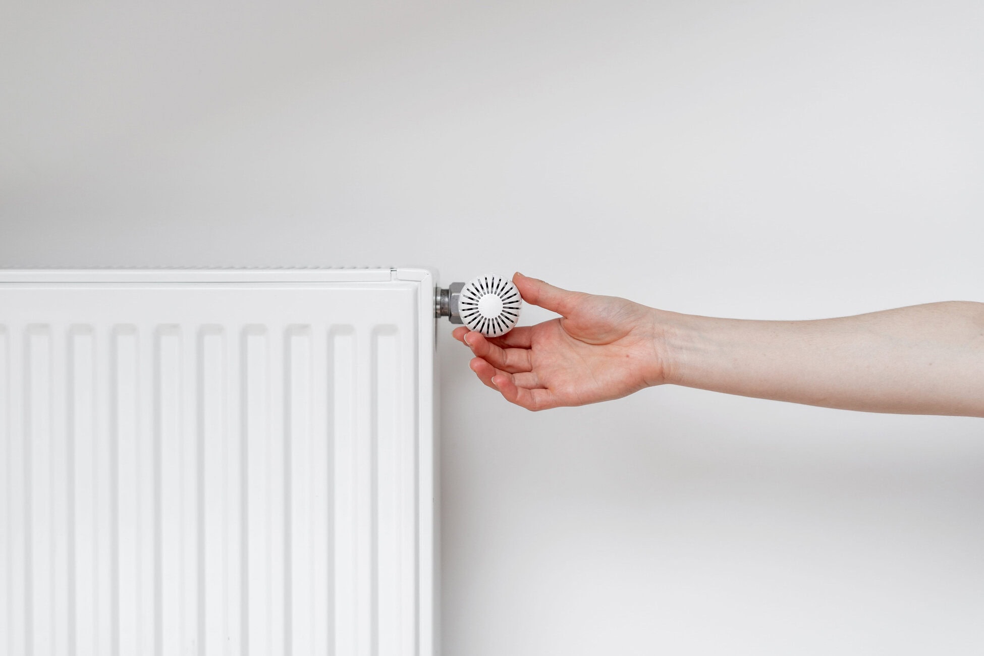 Close-up of a hand turning a circular radiator valve on a white radiator against a pale wall